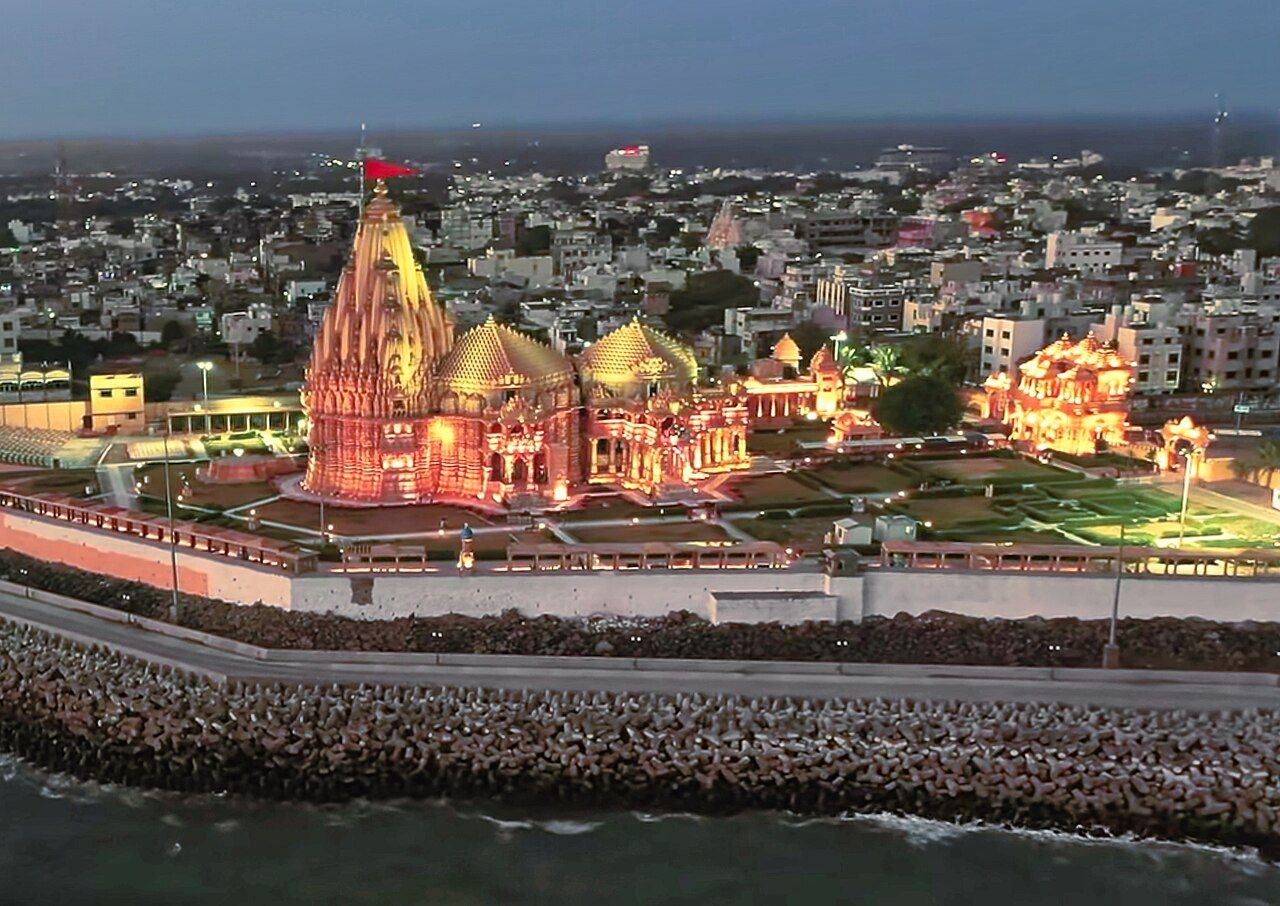 Aerial view of Somnath temple lit at dusk, standing on a sea wall above the Arabian Sea, with the town behind it