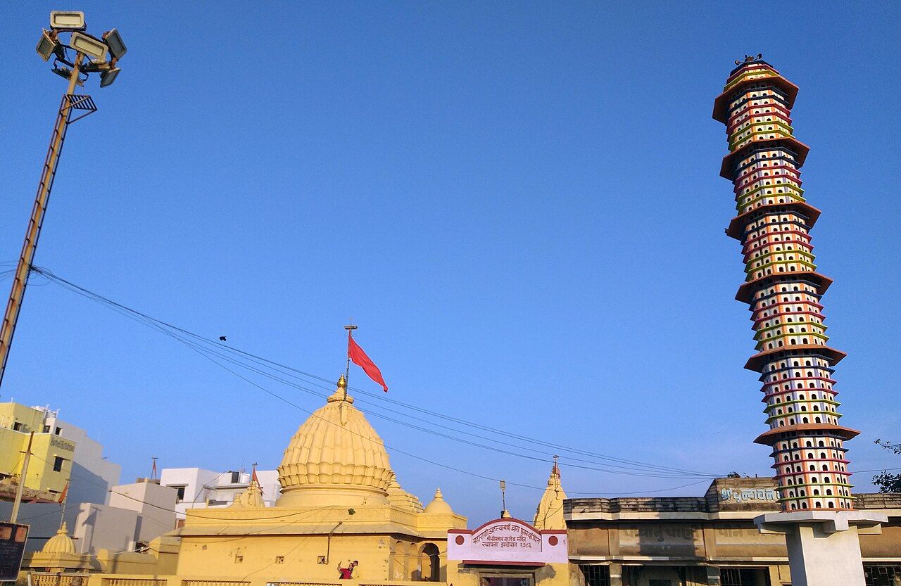 A cream stone temple with a round dome and a red flag, under a blue sky, beside a tall painted tower
