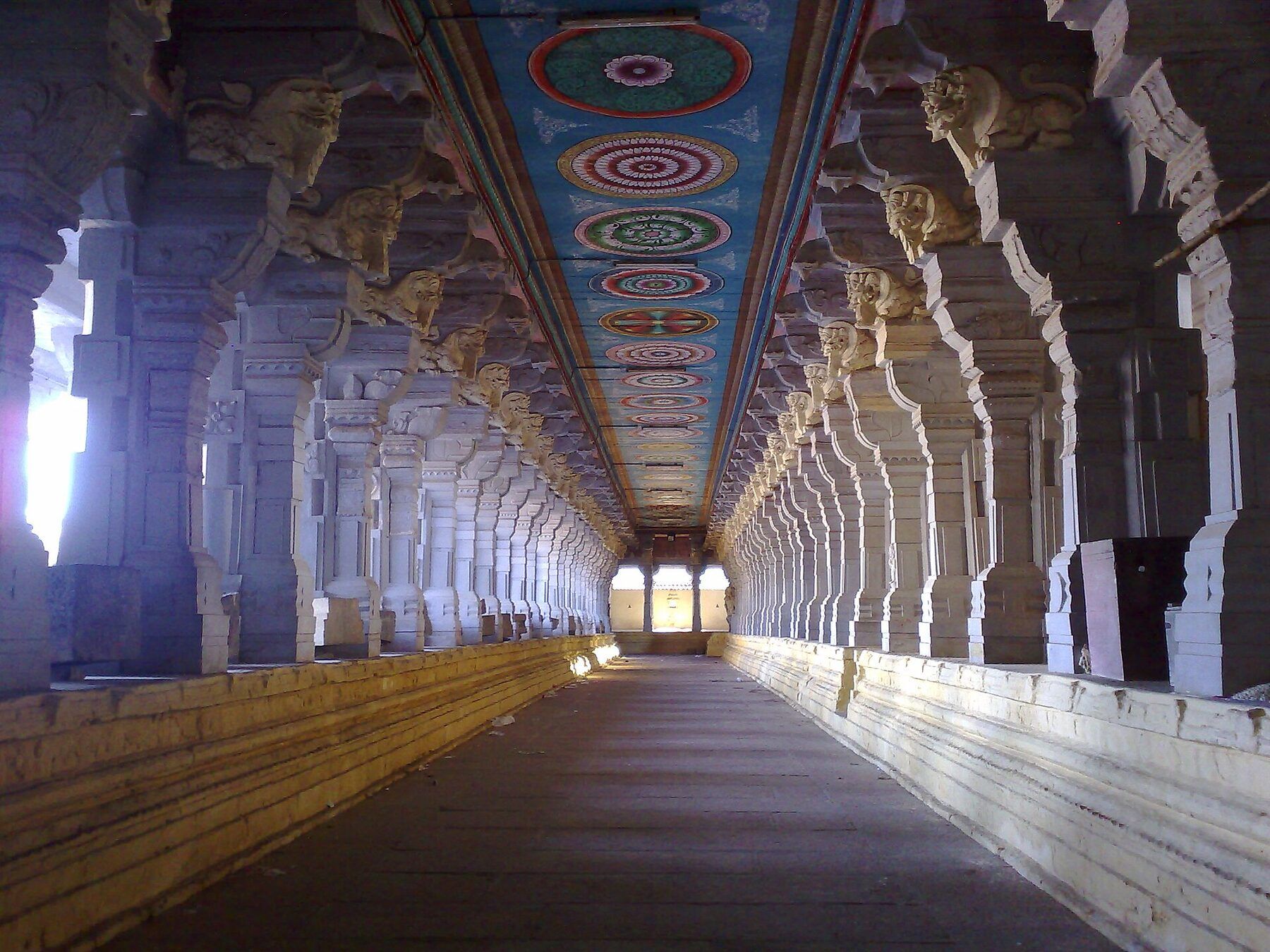 A long, empty stone corridor lined on both sides by carved pillars, with painted circles on the ceiling and daylight at the far end