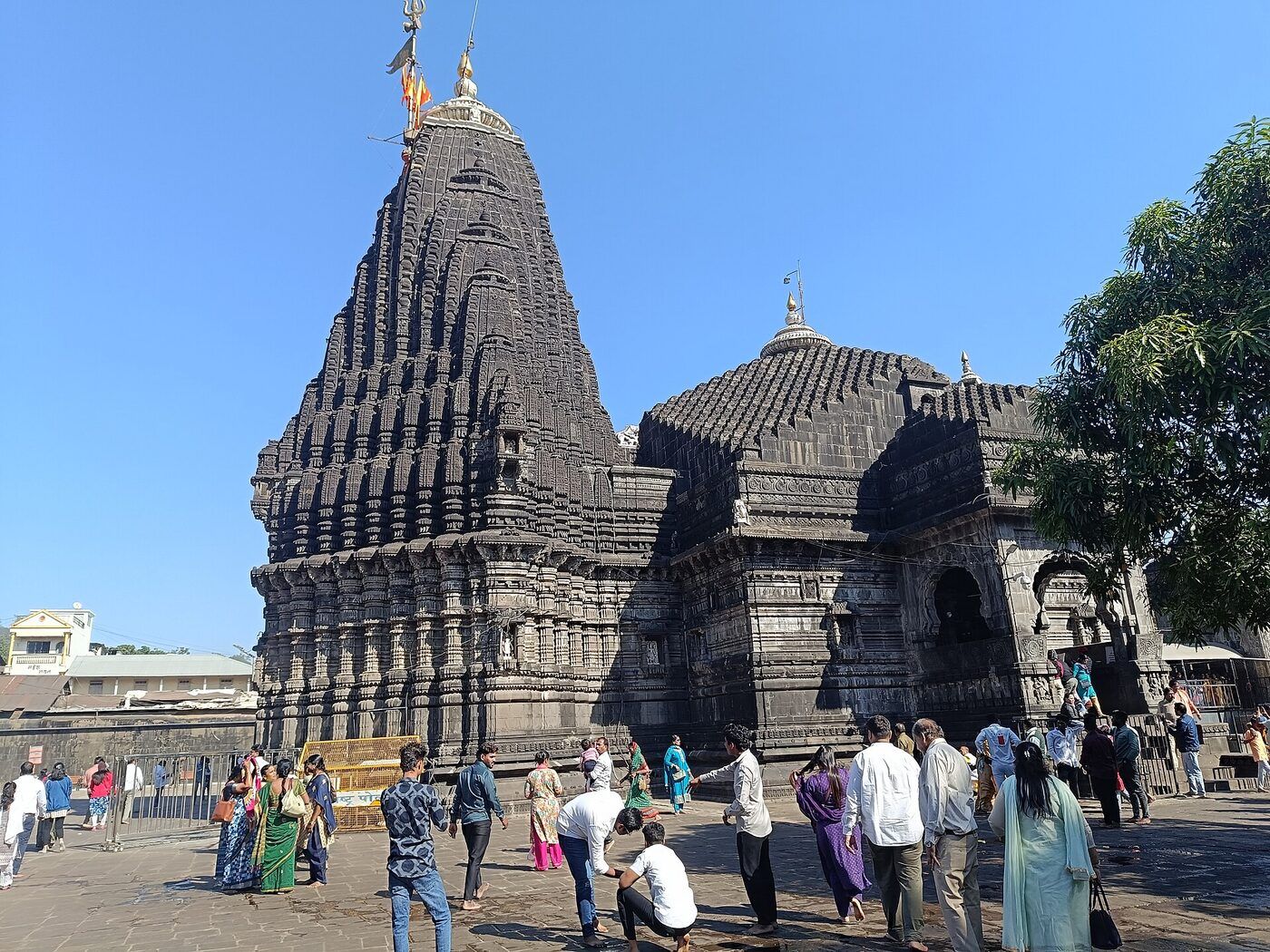 The dark stone temple with its tall ribbed spire, and visitors walking across the paved courtyard in front of it