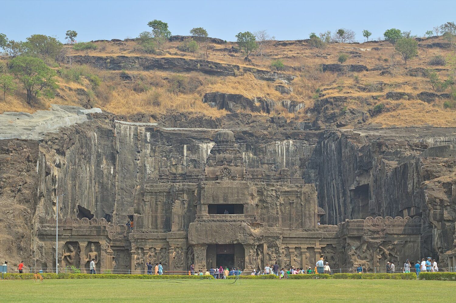 A temple carved out of a dark rock cliff, with a lawn and small groups of visitors in front