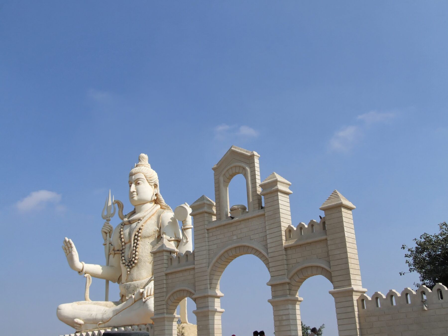 A large white statue of a seated Shiva beside a cream stone gateway with arches, under a blue sky
