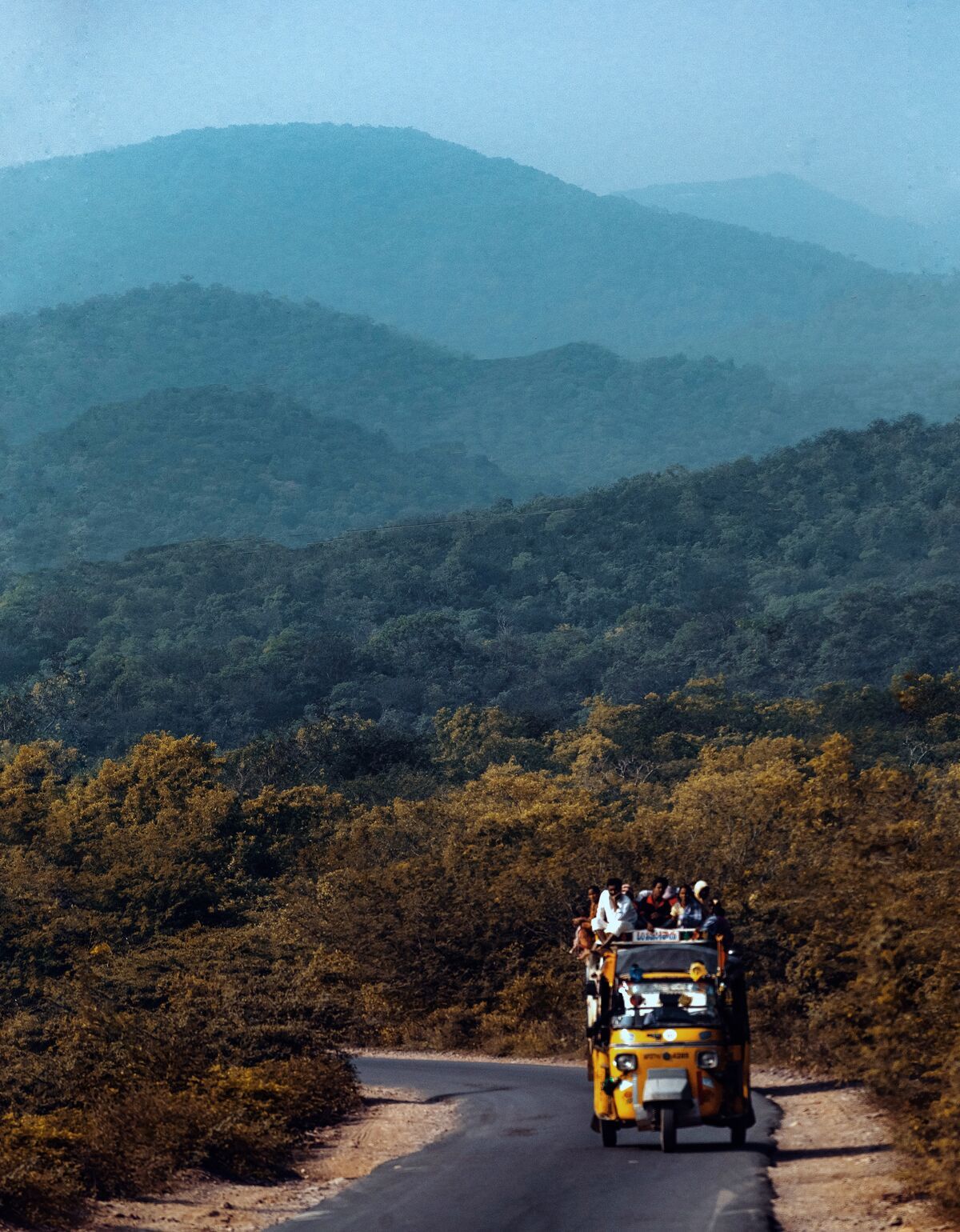 A loaded auto rickshaw on a narrow road through forested hills, with blue ridges behind