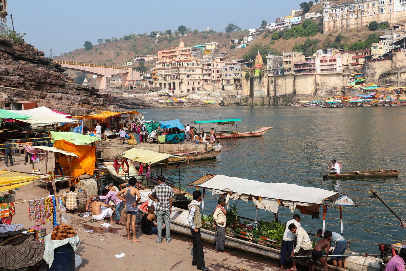 A riverside ghat with stalls and moored boats, and buildings on the hill across the water