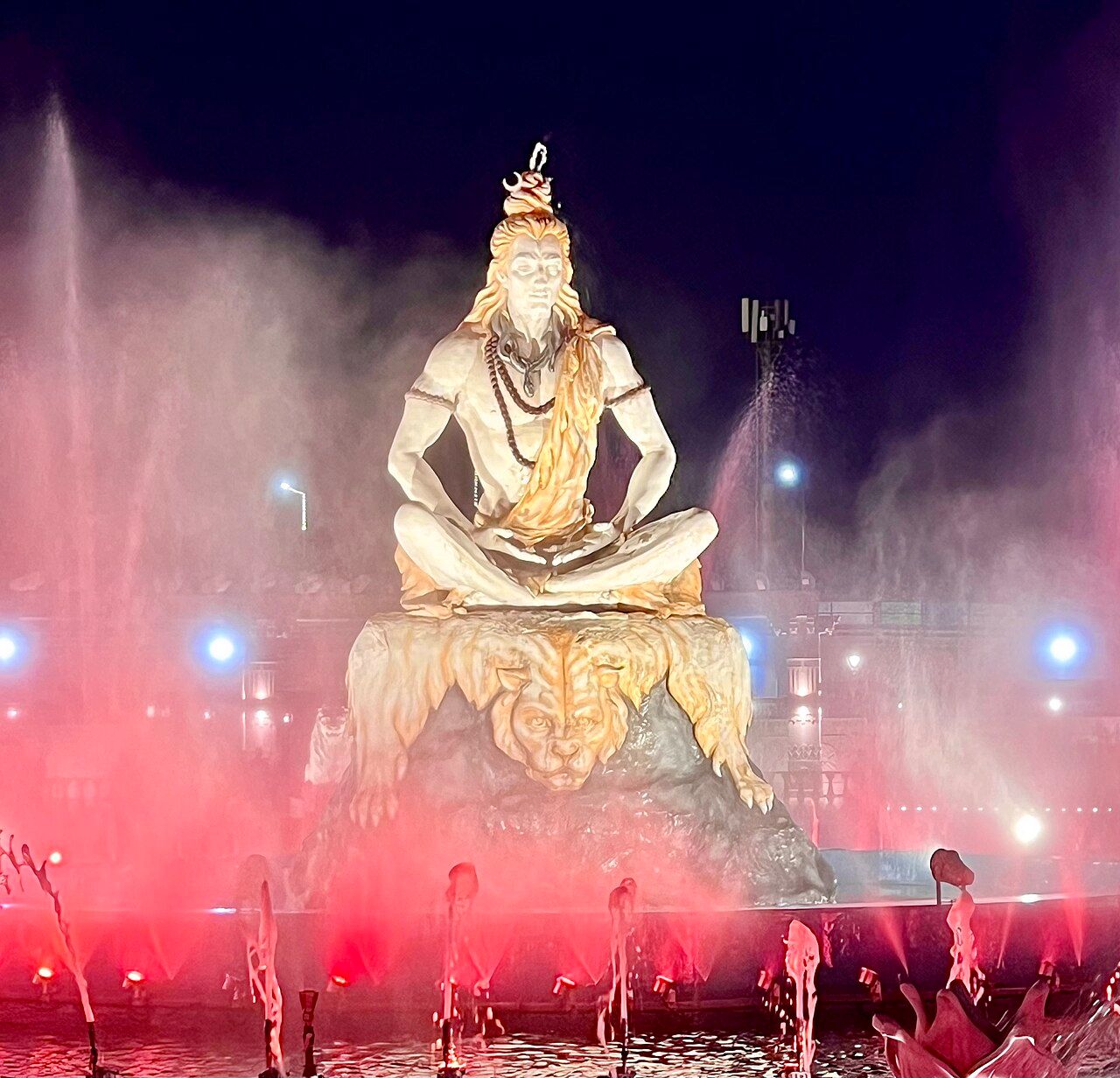 A white statue of Shiva seated on a rock, lit in red and pink, with fountains spraying behind it at night
