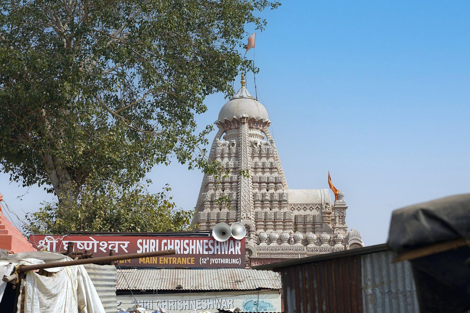 The carved pale spire of the temple rising behind a red sign reading Shri Grishneshwar Main Entrance, with stall roofs in front and a tree to the left