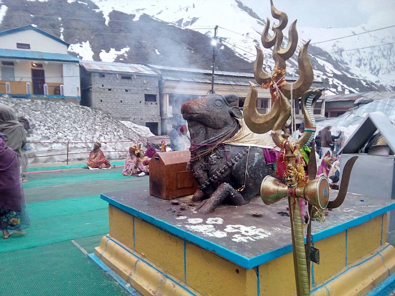 A dark stone Nandi statue on a raised platform with a brass trident beside it, buildings and snow behind