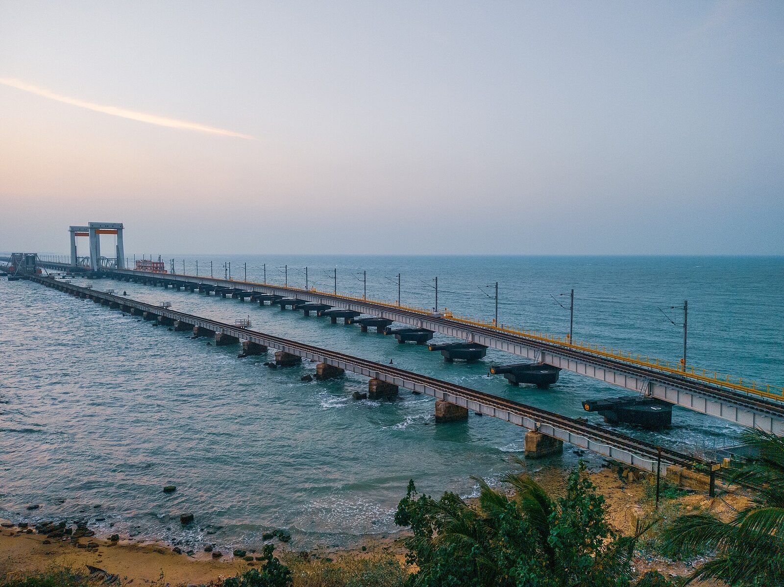 A long railway bridge on low piers crossing blue-green sea, seen from the shore at dusk