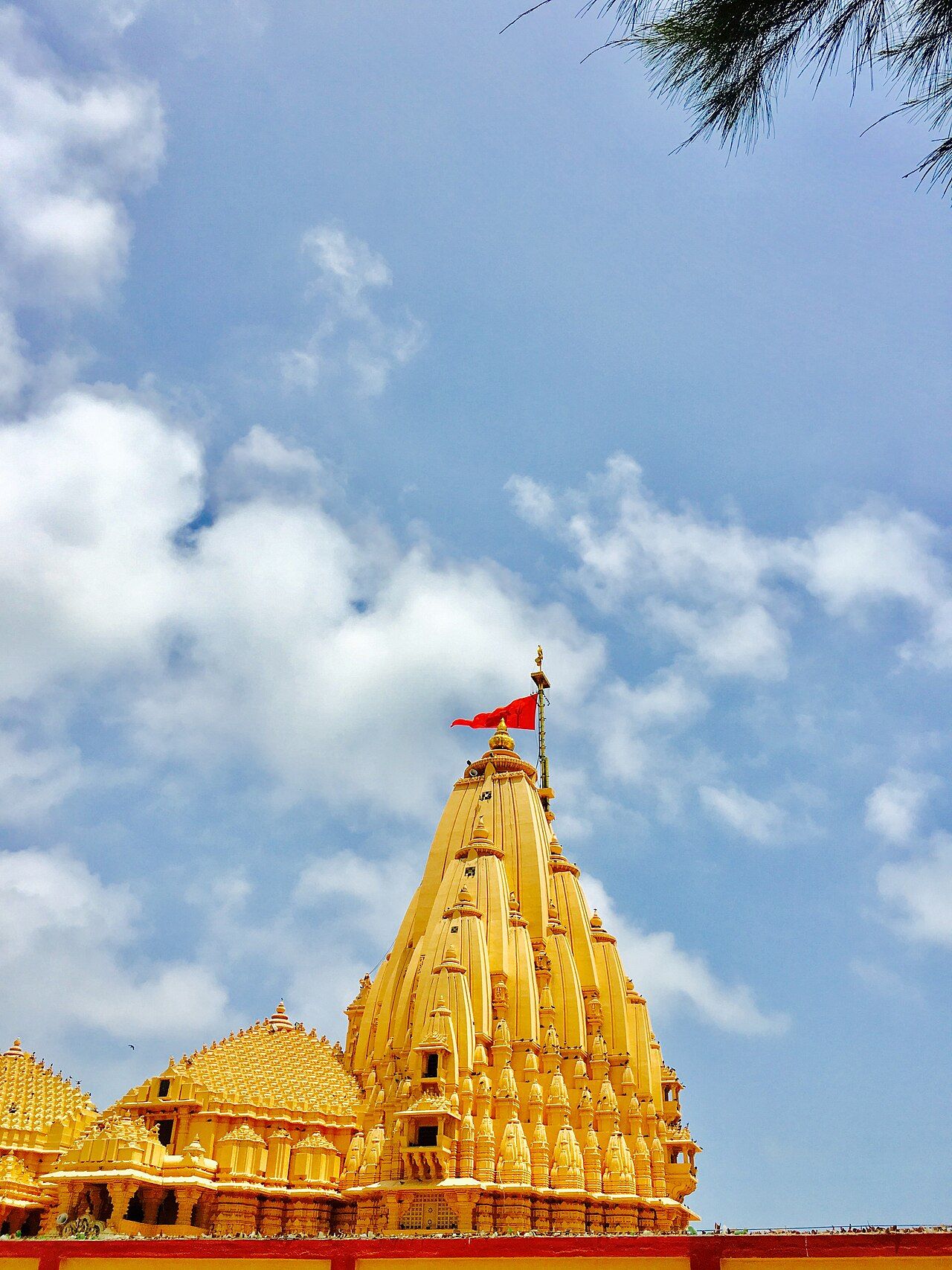 The golden carved spire of Somnath temple with a red flag, against a blue sky with white clouds