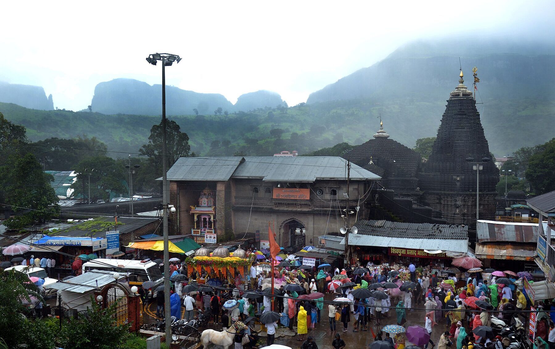 A large crowd with umbrellas and stalls in front of the temple gate building, with the temple spire and misty hills behind