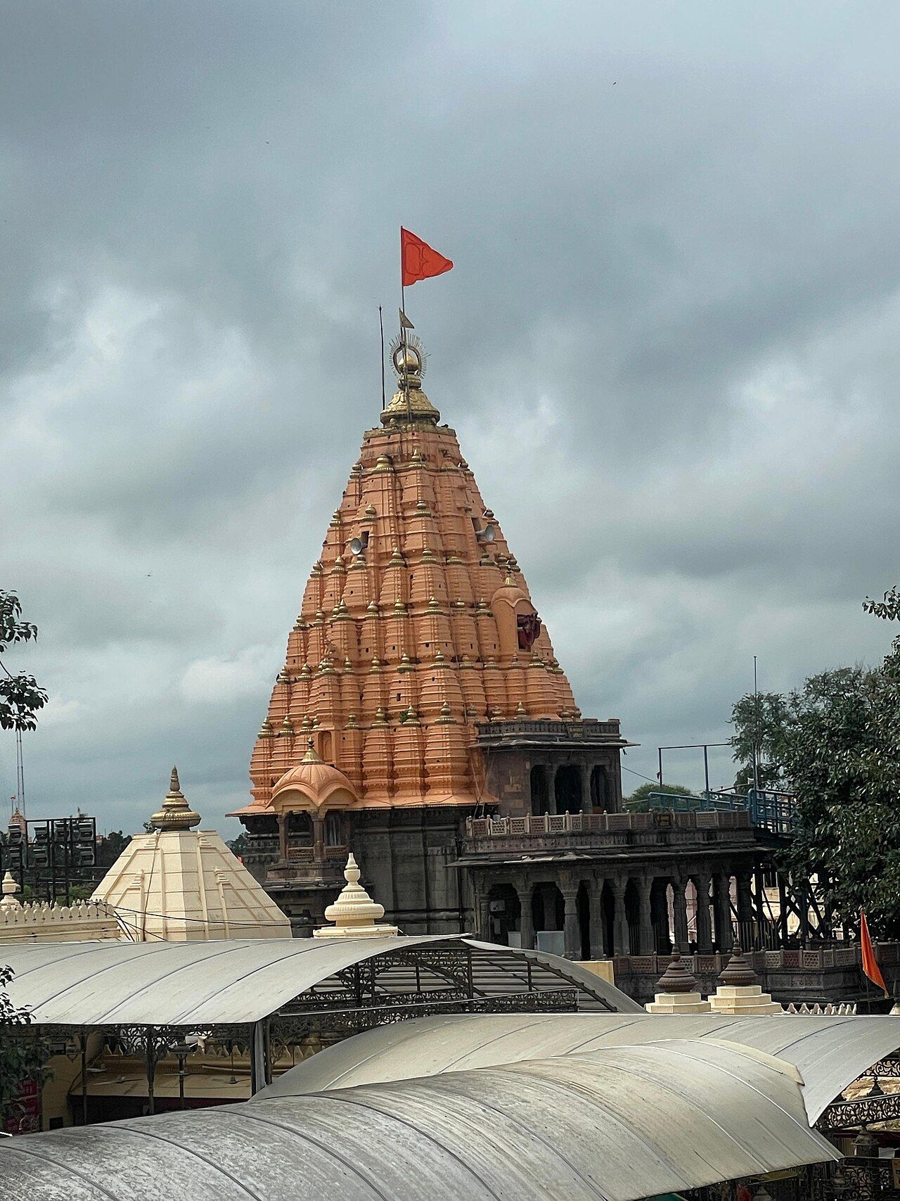 The orange stone spire of Mahakaleshwar temple with a saffron flag, behind curved white canopy roofs, under a cloudy sky
