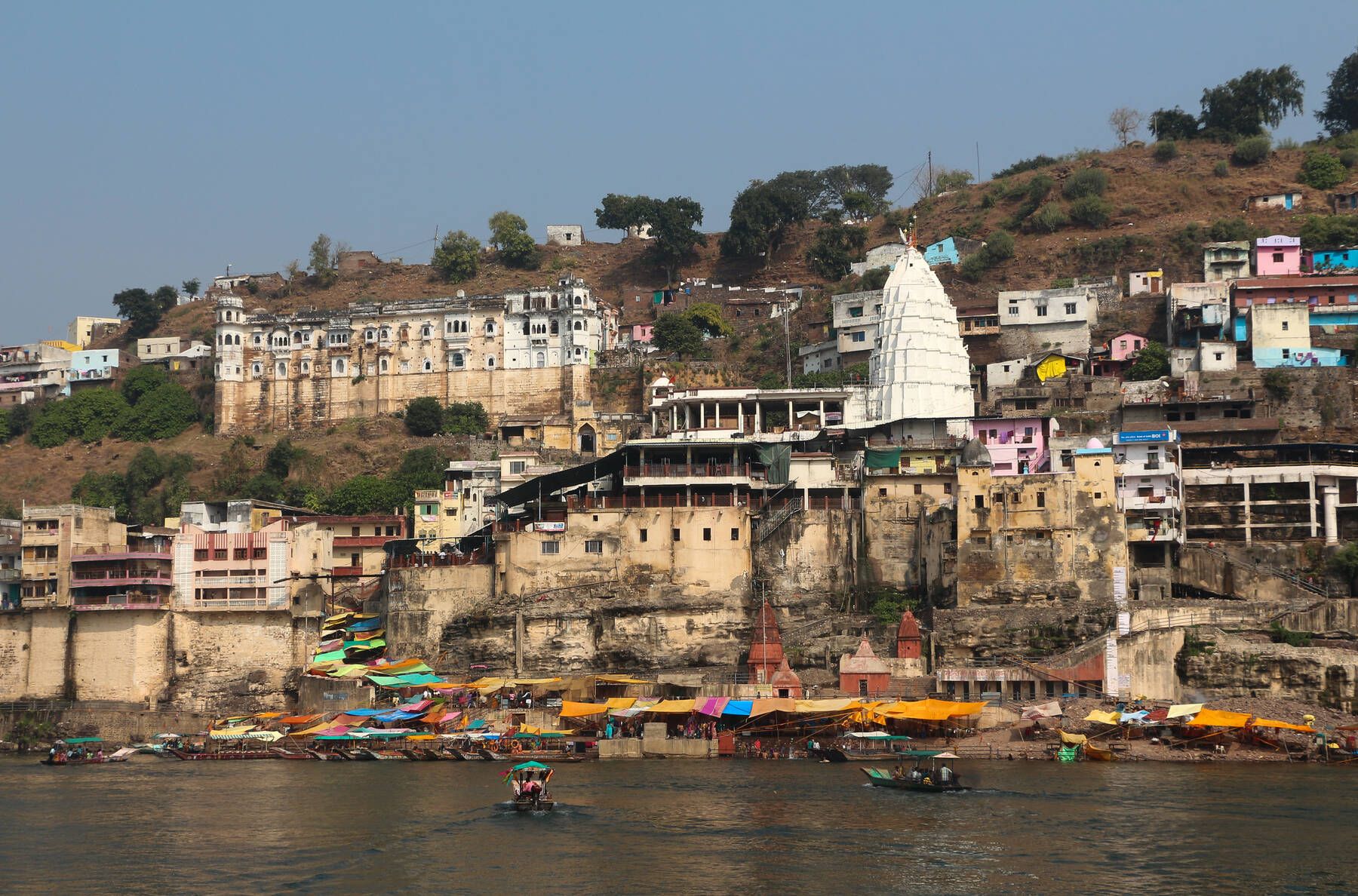 A white temple spire above stone ghats and houses on a hillside, seen across the river