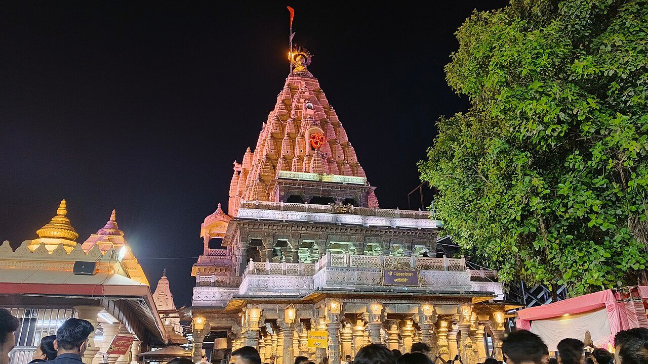 The tall tiered spire of Mahakaleshwar temple lit at night, with a crowd in front and a tree to the right