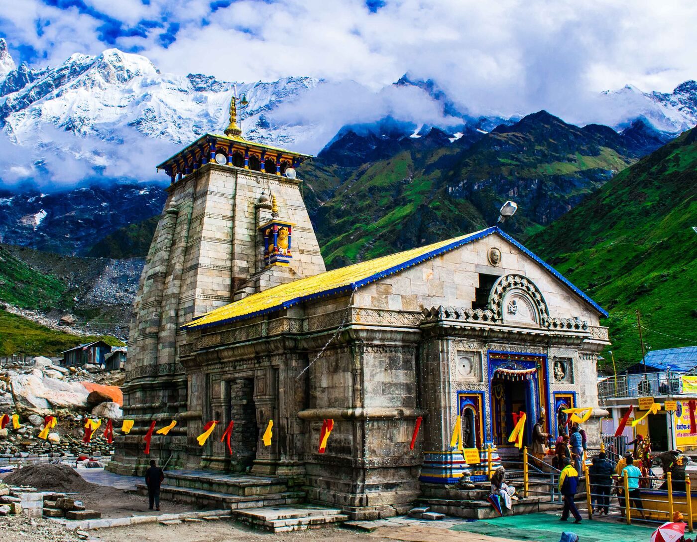 Kedarnath temple in front of green slopes, with snow-capped peaks and cloud behind