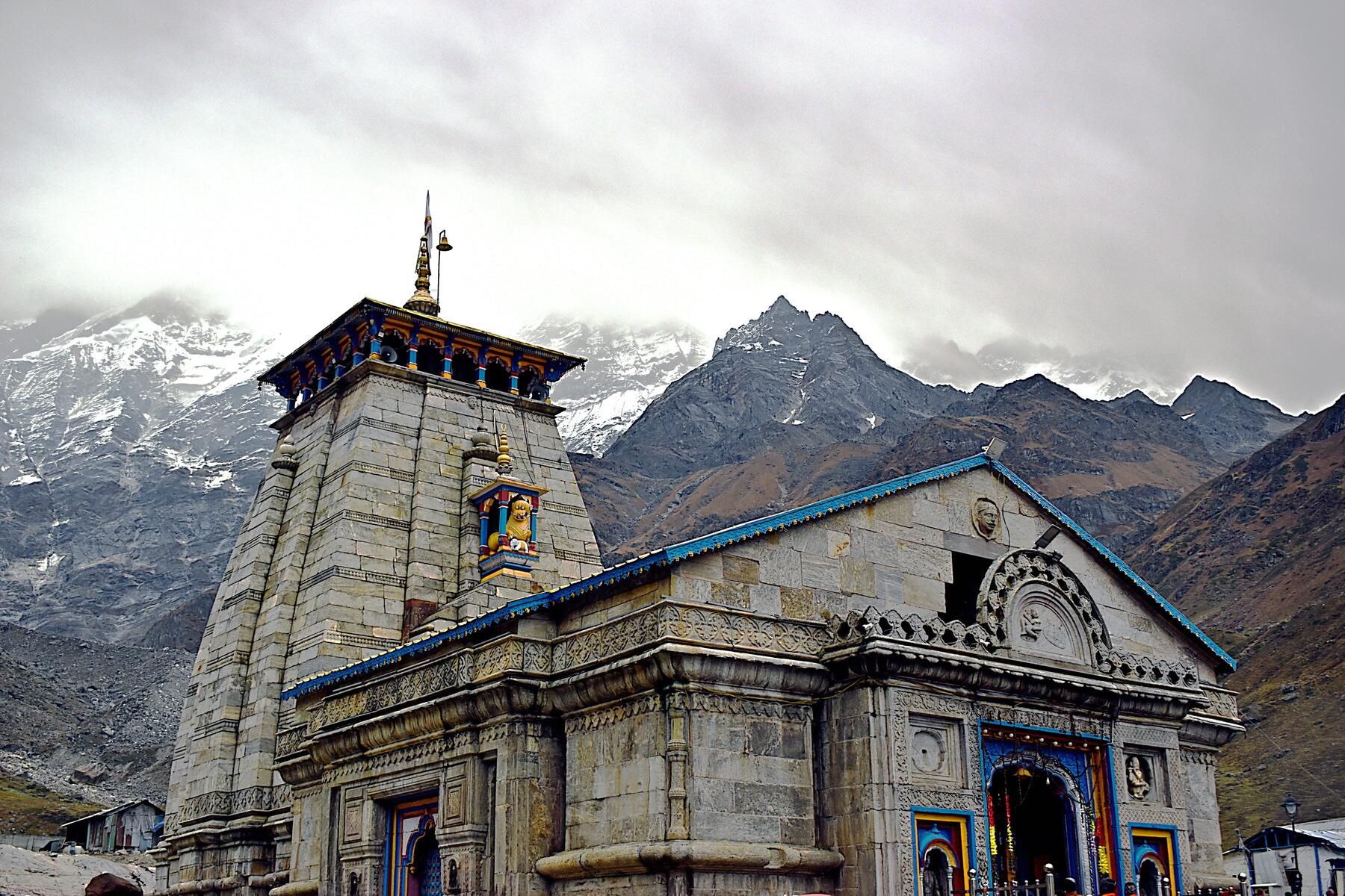 Kedarnath temple, built of grey stone with a tall spire, with snow-streaked peaks behind it under cloud